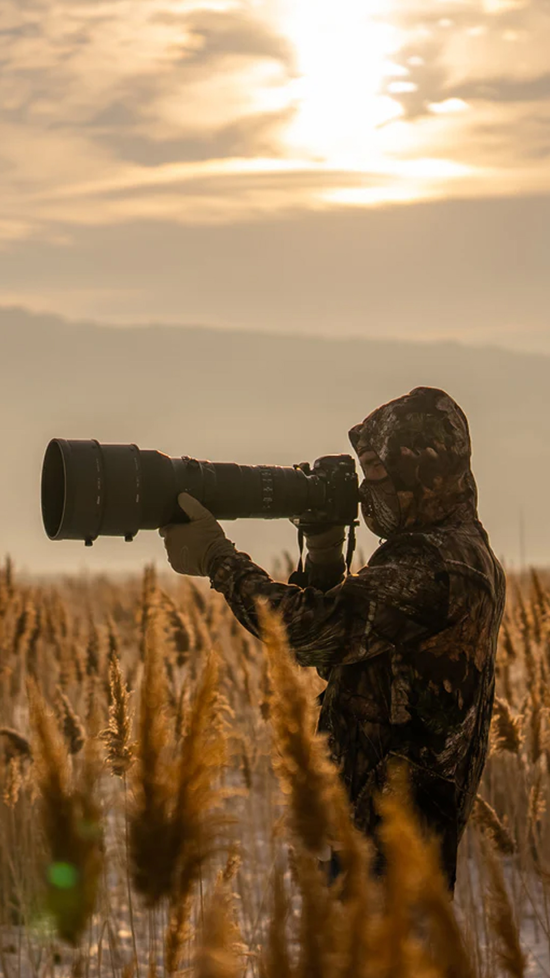 Photographer, Artem Shestakov, dressed in camouflage gear is aiming a large telephoto lens in a field at sunset, capturing a serene natural scene.