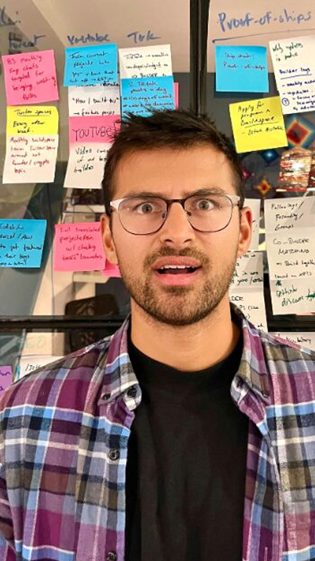 Farza in glasses and a plaid shirt, standing in front of a glass wall filled with colorful sticky notes; he appears surprised or excited.