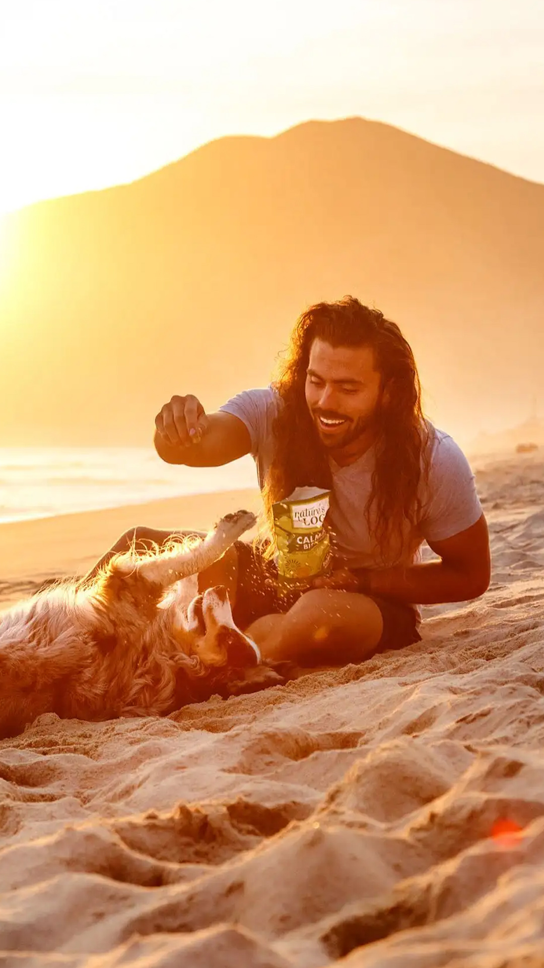 Tristan Hamm with long hair playing with his dog on a sandy beach during sunset, holding a bag of pet treats labeled 'Nature's Logic.'