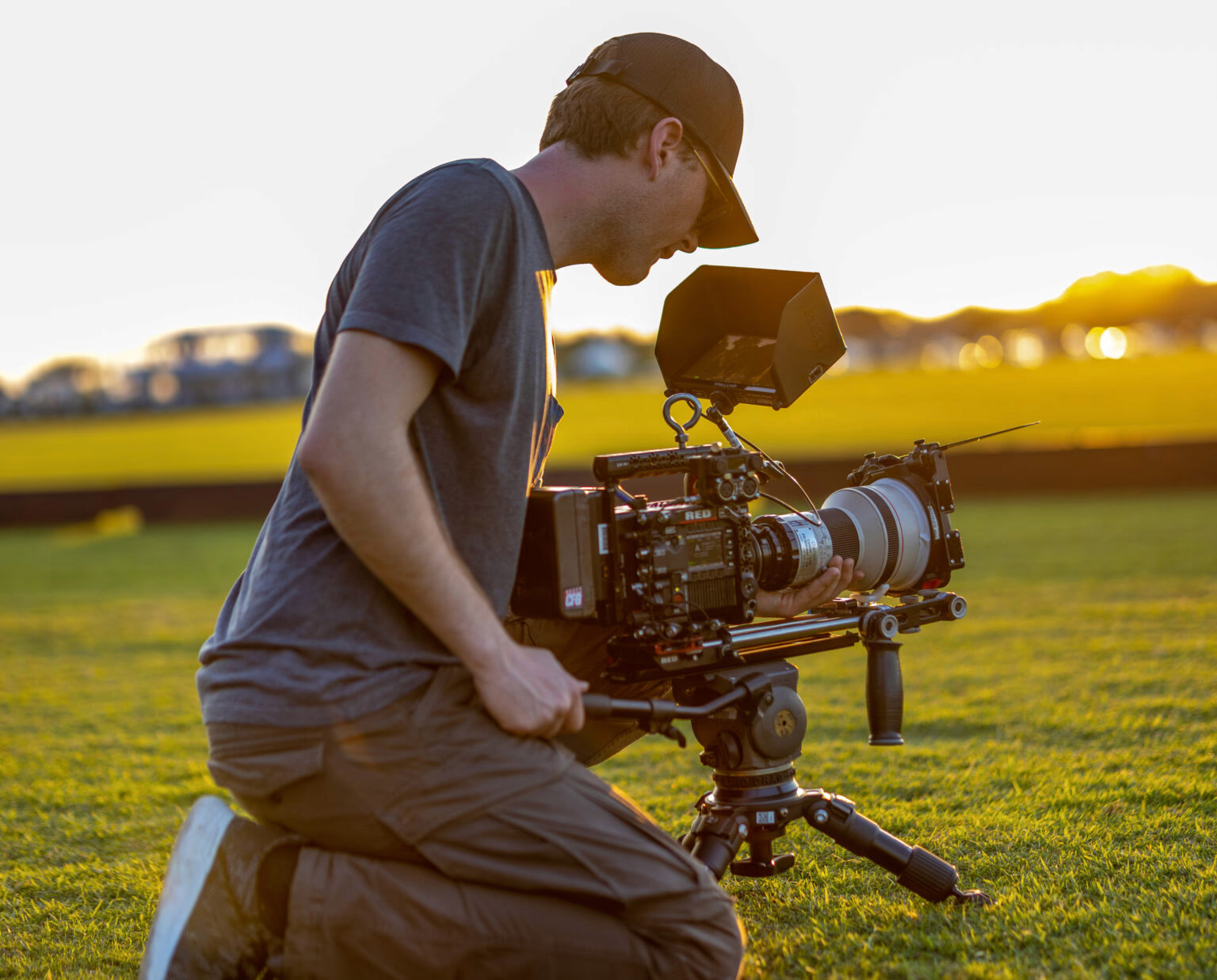 Filmmaker, Quinn Halleck, kneeling with a professional video camera on a grassy field during golden hour, focused on capturing footage.