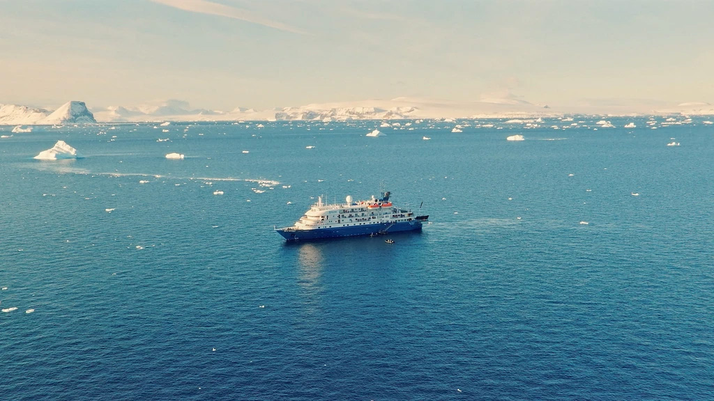 A cruise ship navigating through icy Antartic waters with scattered icebergs, against a backdrop of snowy mountains.