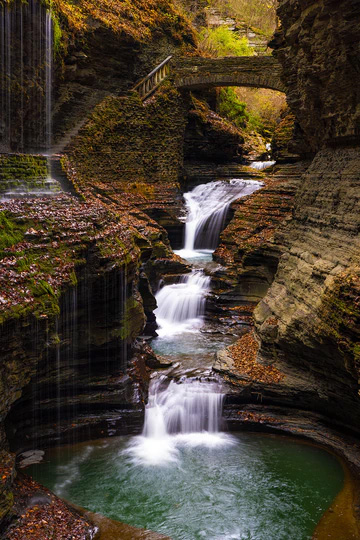 Scenic waterfall cascading through layered rocks with a stone bridge overhead, surrounded by lush greenery.