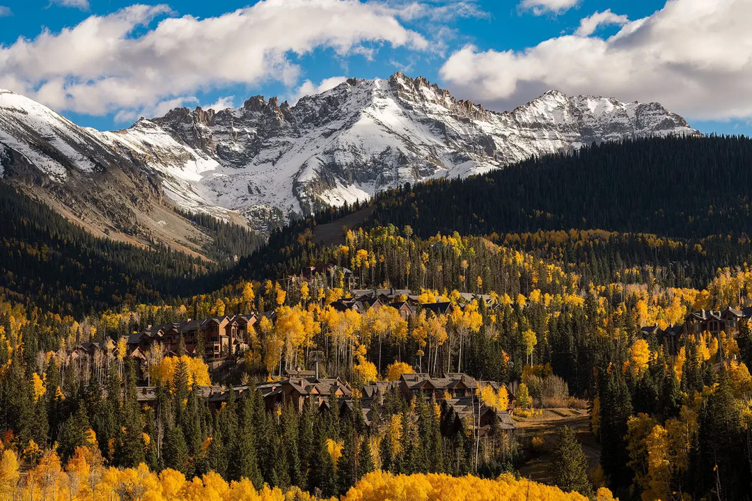 Autumn landscape with yellow aspens and evergreen trees leading up to a snow-capped mountain range under a partly cloudy sky.