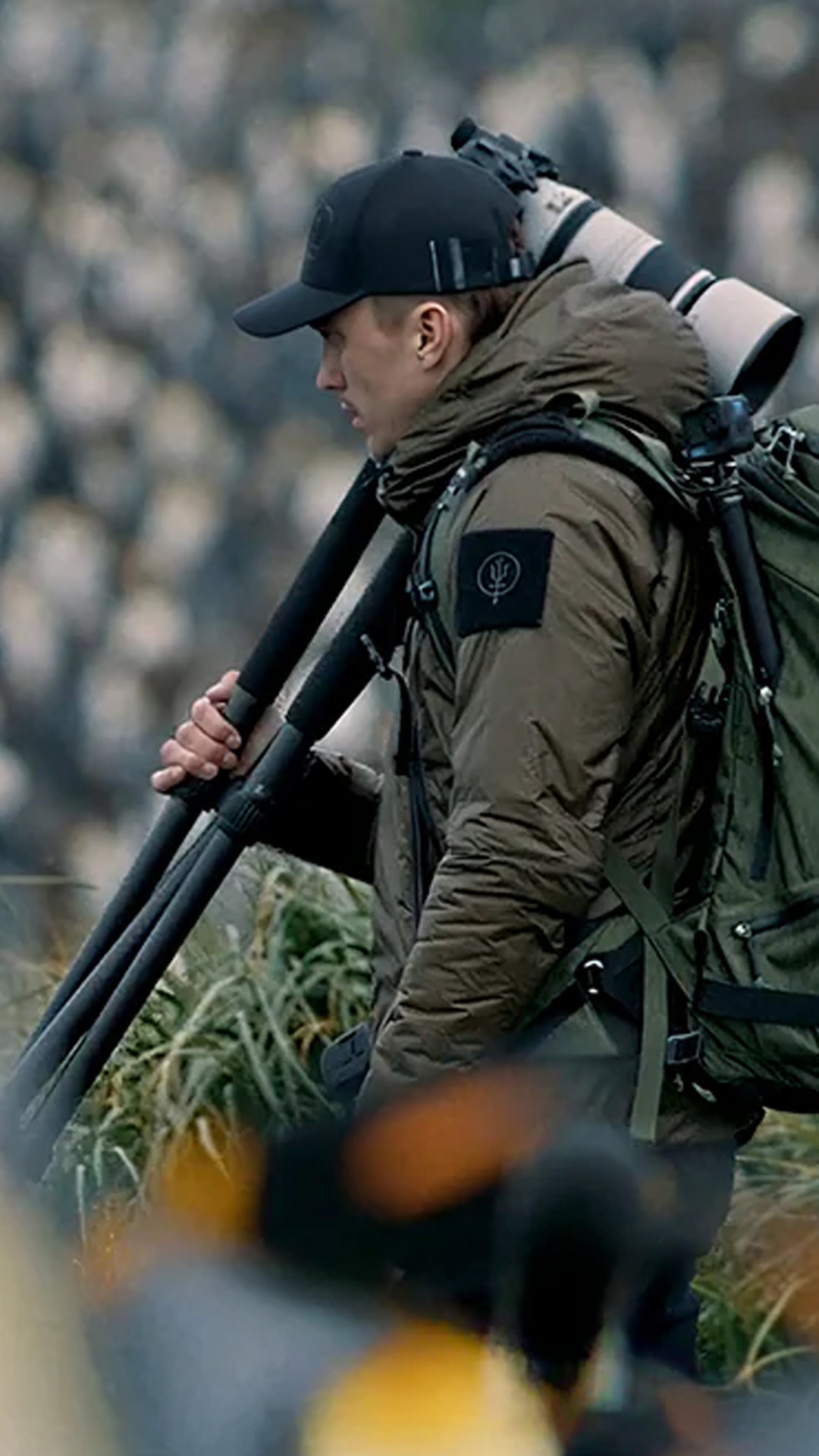 Photographer, Artem Shestakov, carrying a large camera and tripod, dressed warmly and surrounded by a blurred backdrop, likely of penguins.