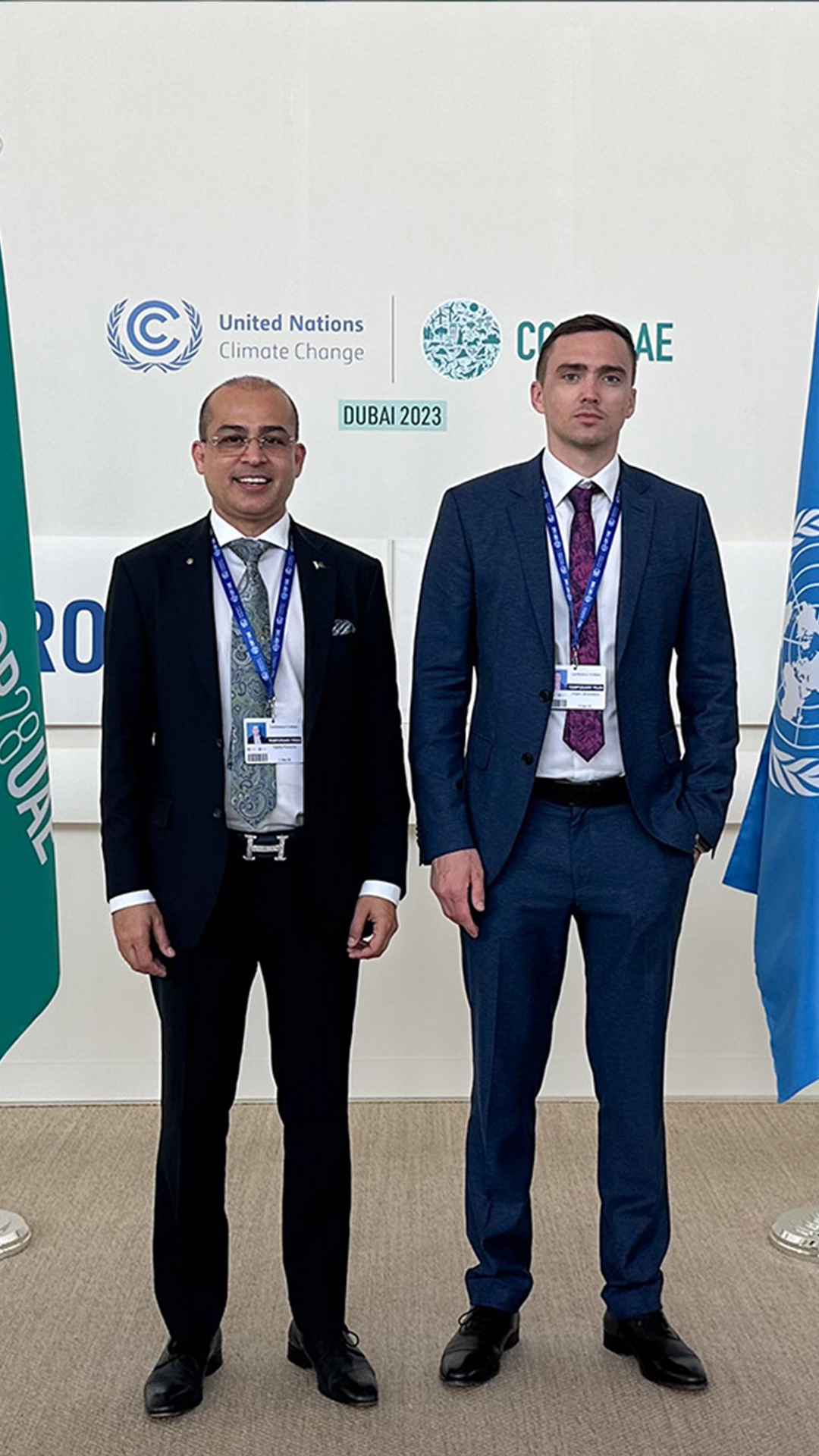 Two men dressed formally, standing together at a UN Climate Change event in Dubai, with event branding in the background.