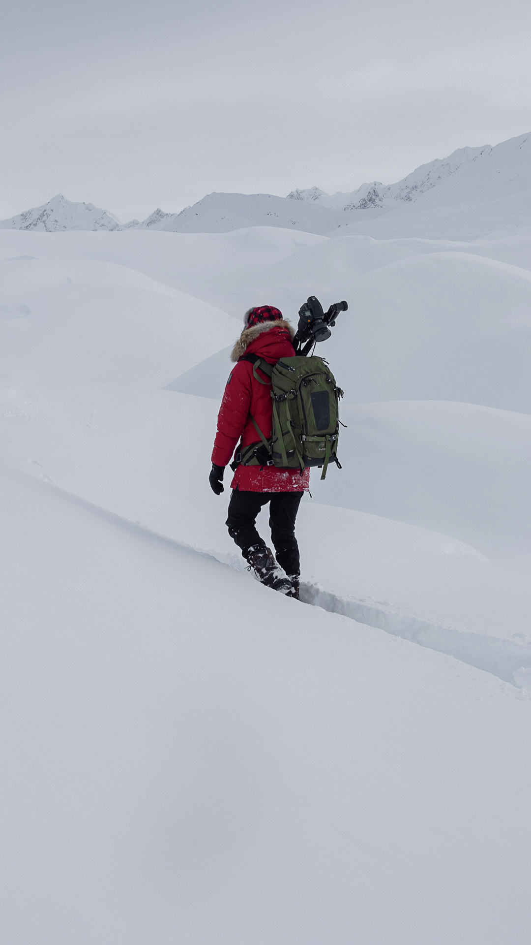 Artem Shestakov, trekking through deep snow in a mountainous, snowy landscape, carrying a large backpack and camera gear.