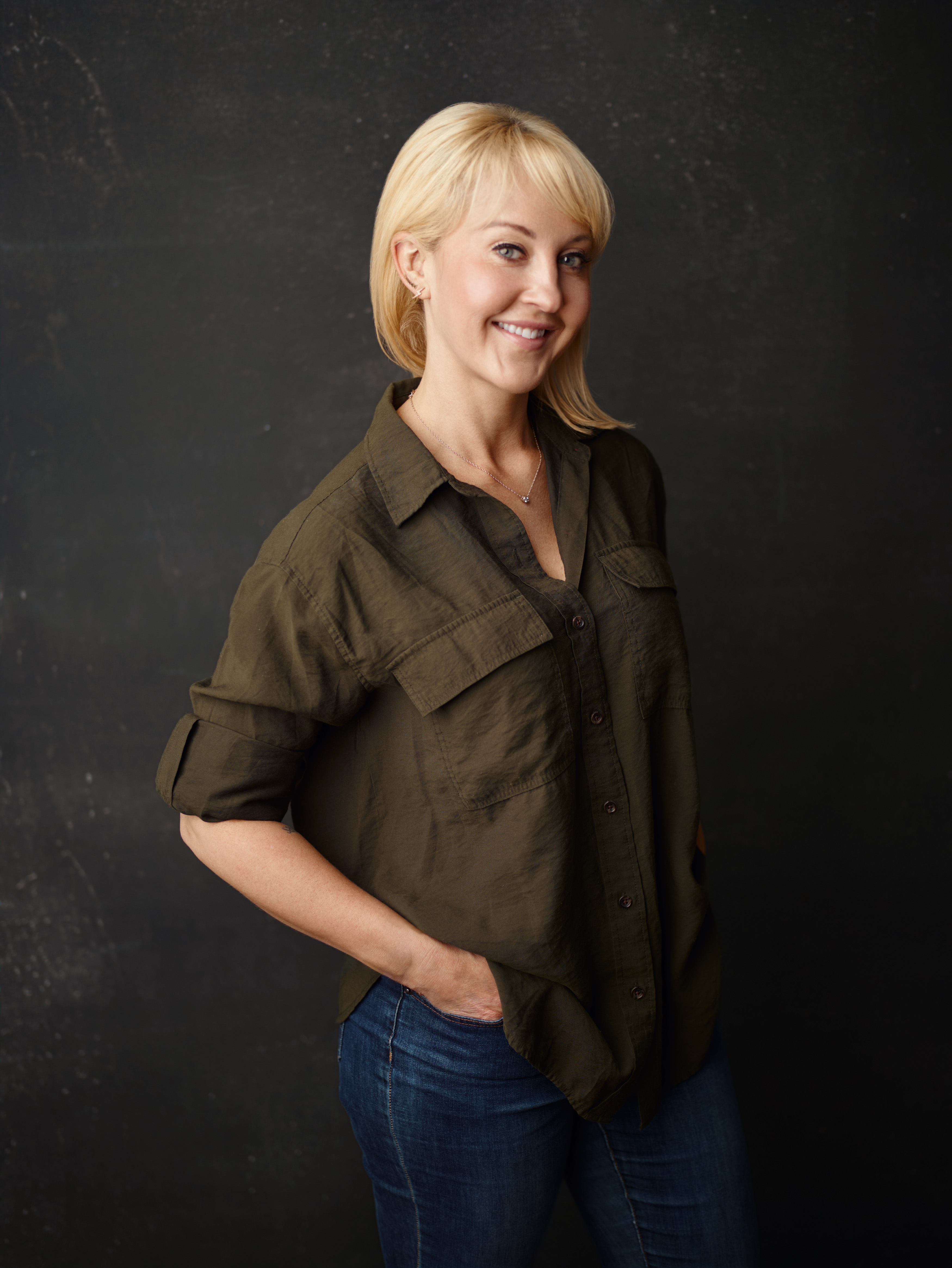 Molly Shelestak, with short blonde hair wearing a loose olive green button-up shirt and jeans, standing against a dark background