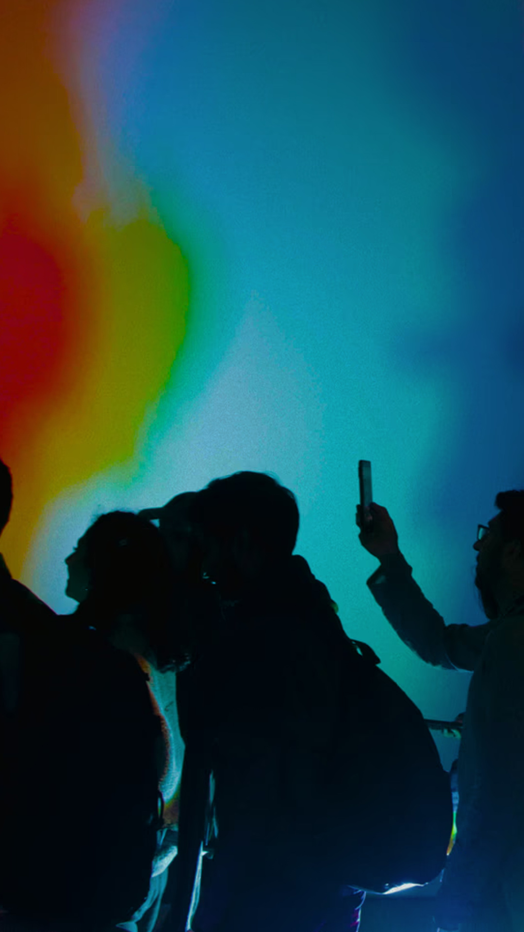 Group of people standing in front of a multicolored, gradient-lit wall; one person holds a smartphone up, presumably taking a photo.