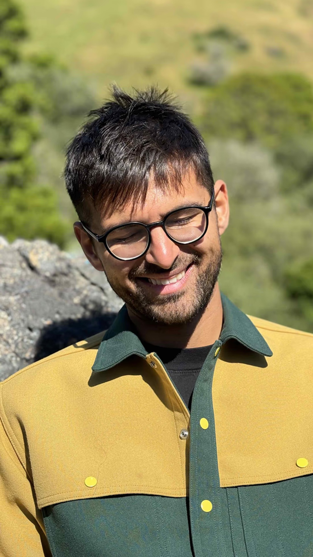 Farza wearing glasses and a green and yellow jacket, smiling with eyes closed while outdoors, with greenery in the blurred background.