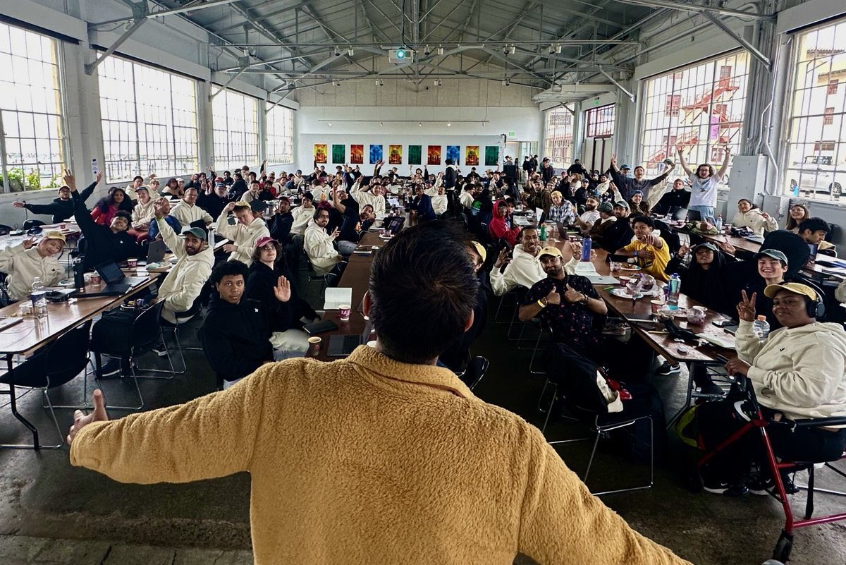 Large group of builders sitting at tables in a spacious hall for buildspace, facing the camera, with Farza in the foreground taking a photo from the back; the atmosphere is lively.