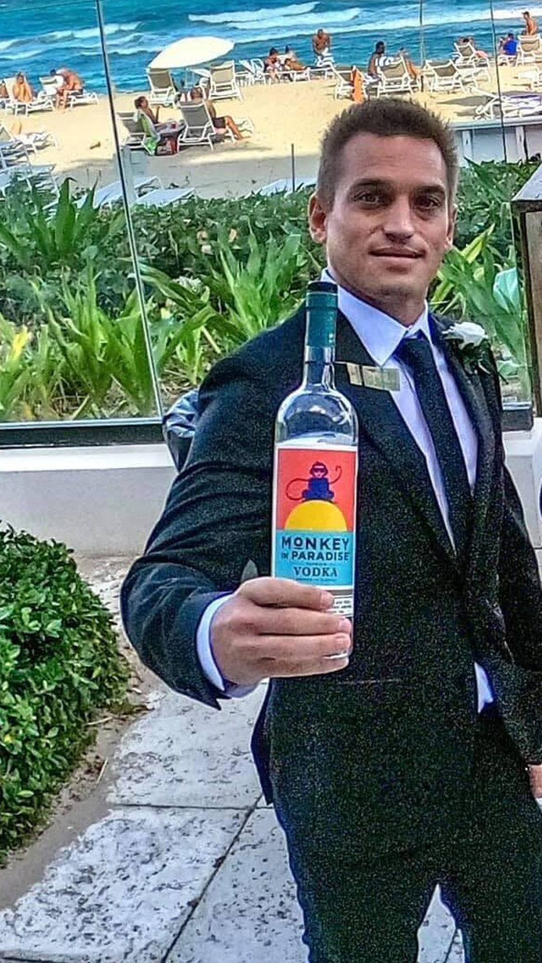 Co-Founder Alex Kowtun holding a bottle of 'Monkey in Paradise Vodka' with a beach scene in the background, people lounging on chairs near the ocean.