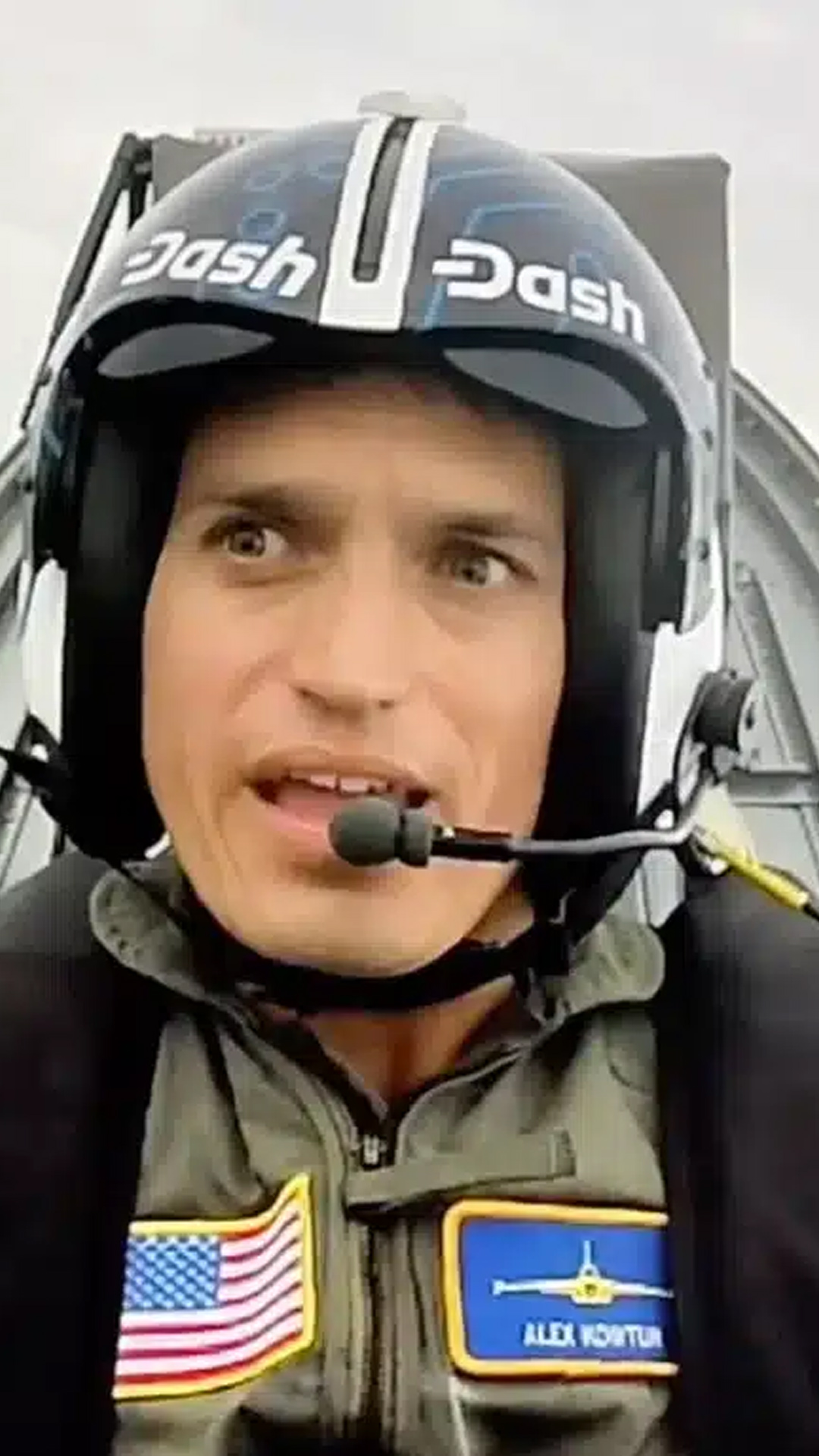 Close-up of pilot, Alex Kowtun, wearing a helmet labeled 'Dash' and speaking into a headset microphone, inside a cockpit.