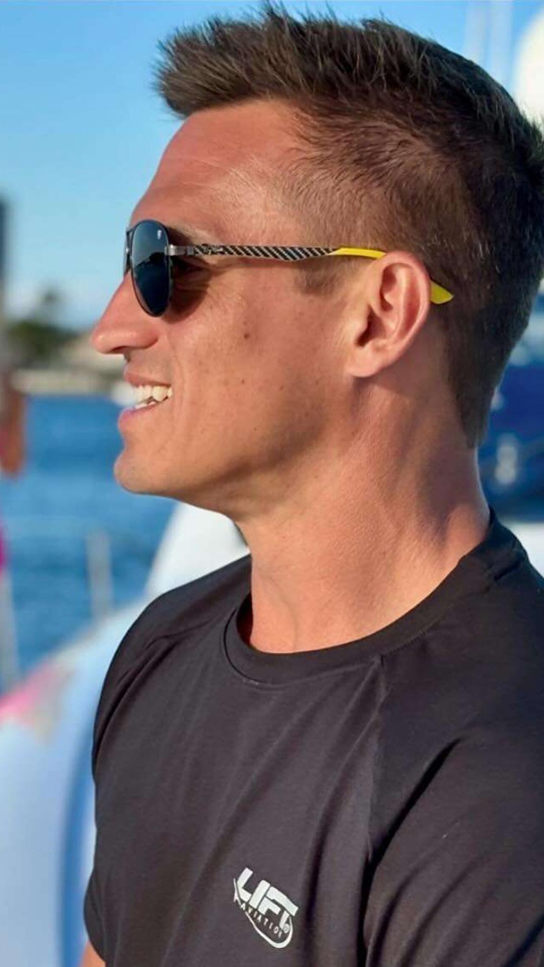 Side profile of Alex Kowtun in sunglasses and a black t-shirt with 'Lift' logo, enjoying a sunny day on a boat with water in the background.