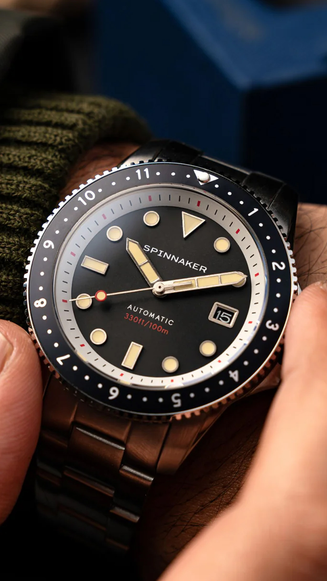 Close-up of a Spinnaker brand automatic wristwatch with a black dial, illuminated markers, and a bezel showing minute markers; the watch is worn on the wrist.