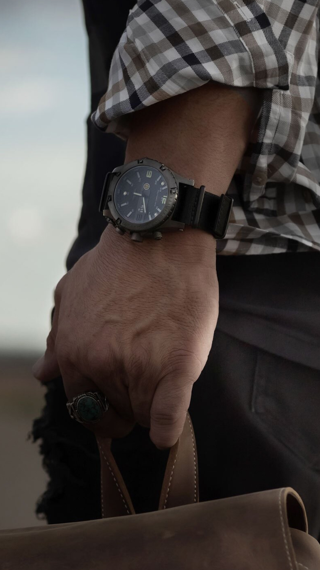 Hand wearing a rugged watch, holding a leather bag; the person is dressed in a plaid shirt, visible in natural lighting with a blurred background.