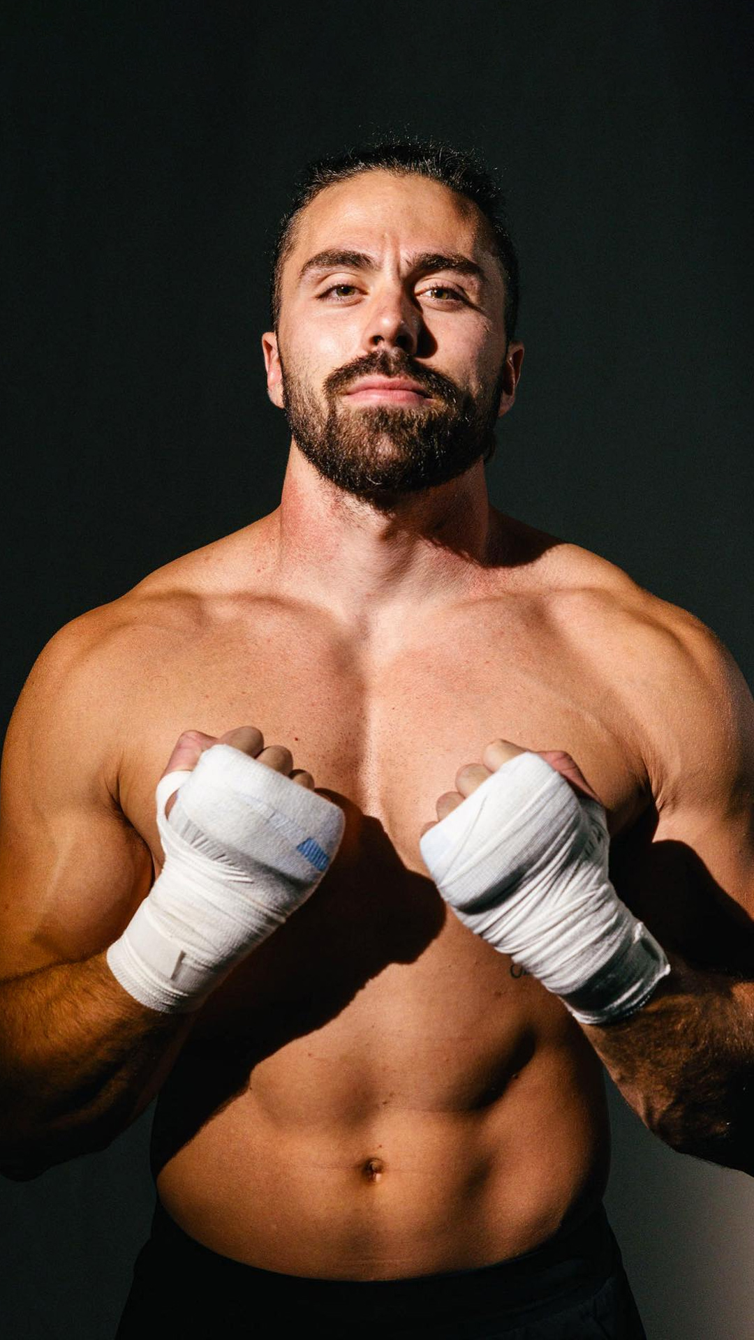 Professional boxer, Tristan Hamm, with a beard, shirtless, with fists raised and wrapped in white bandages, standing against a dark background.