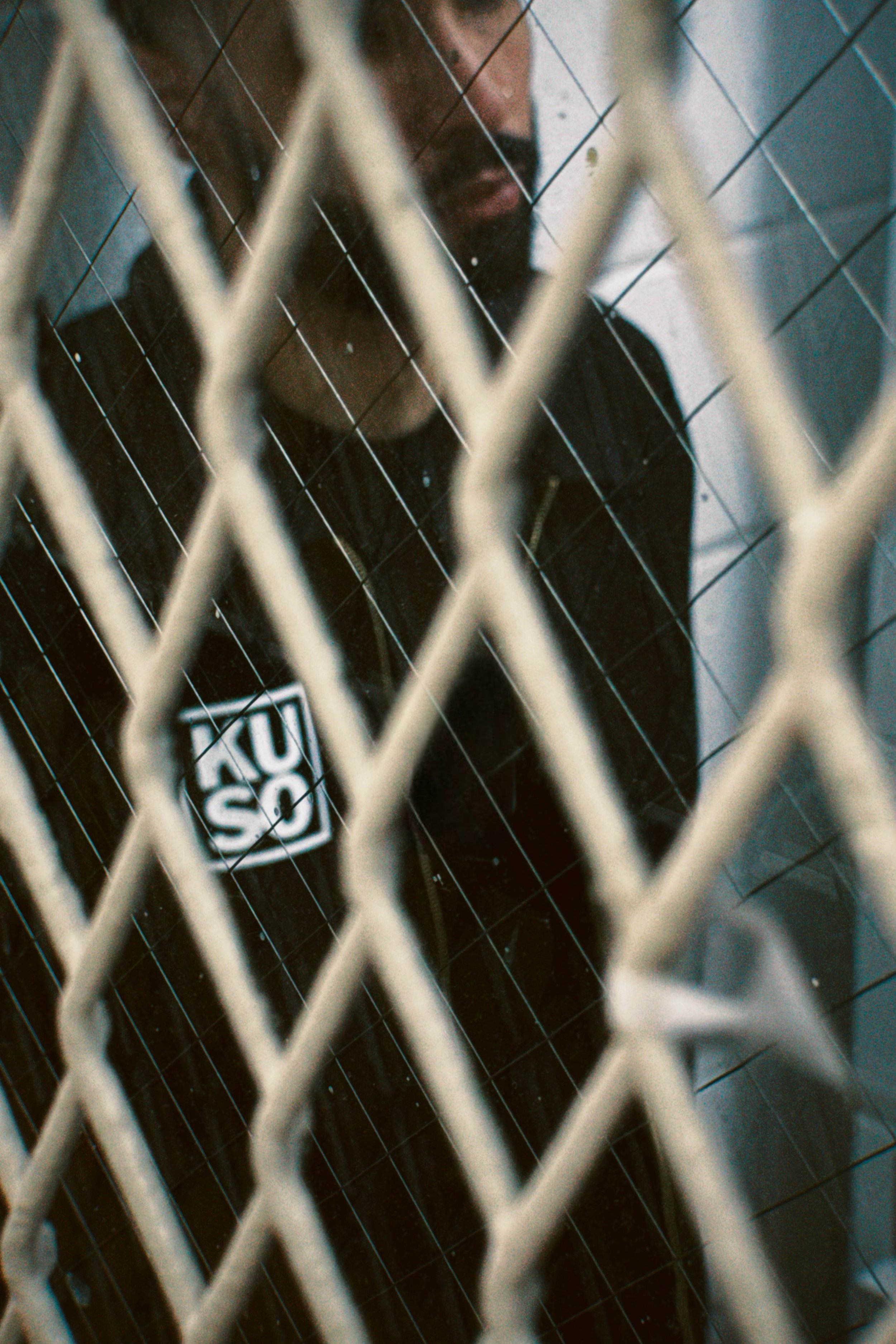 Blurred figure of a man behind a metal fence, partially visible with a reflective surface; he wears a shirt with the 'Kuso' logo.