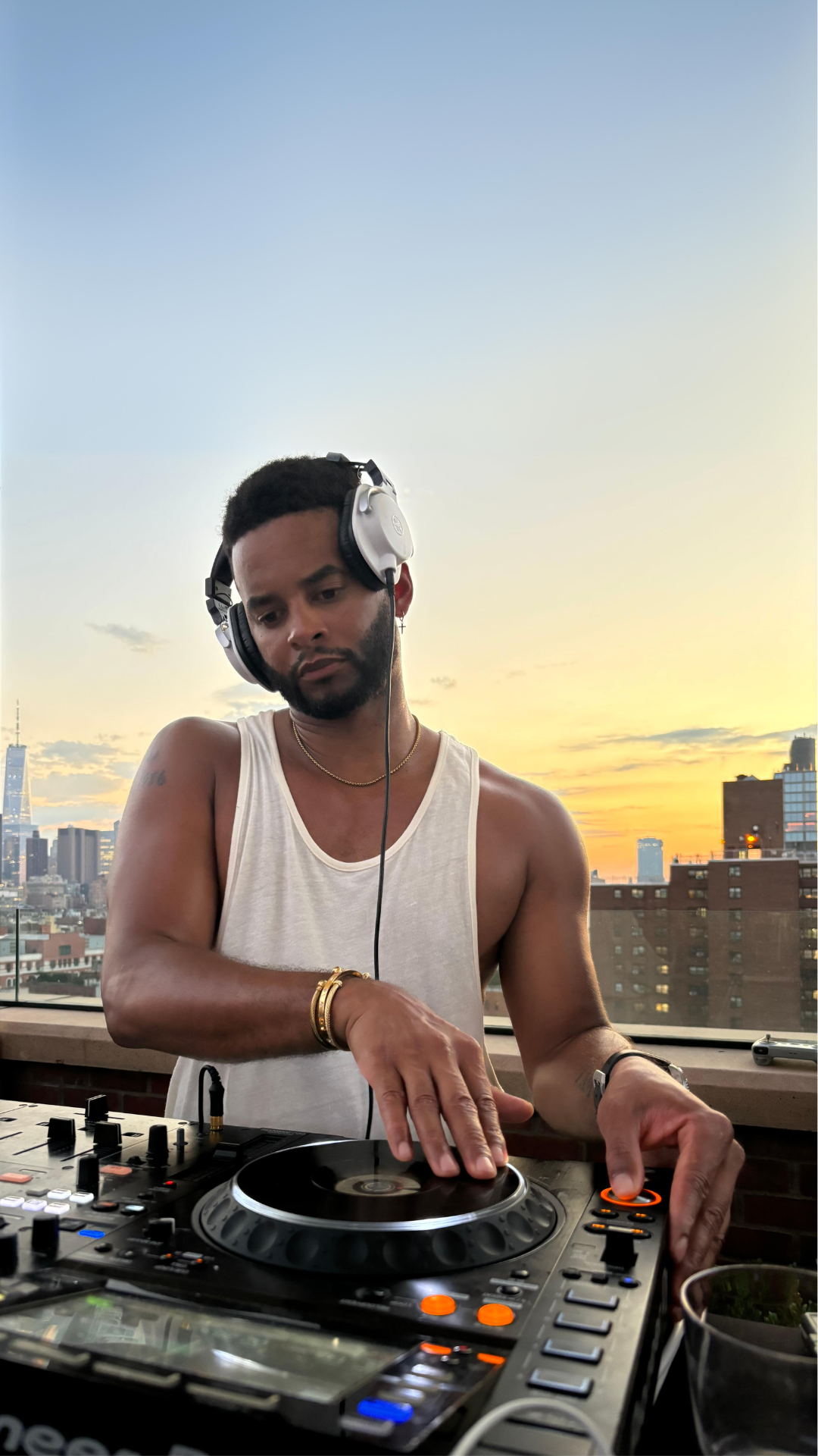 DJ Mike Press wearing a white tank top and headphones, playing music on a console with a cityscape and sunset in the background.