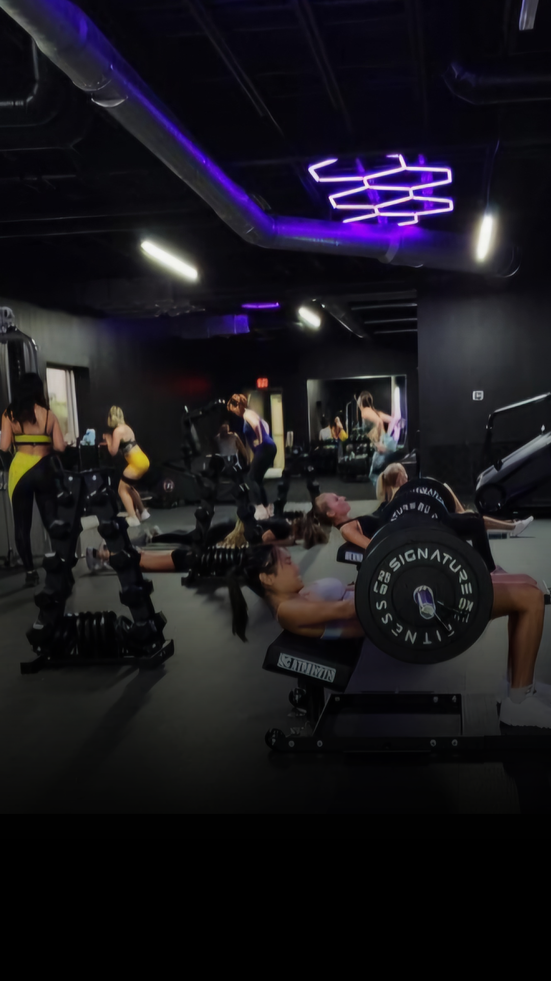 A scene inside a gym with a group of women performing exercises, including hip thrusts with barbells, with signature lighting and gym equipment surrounding them, conveying a focused workout session.