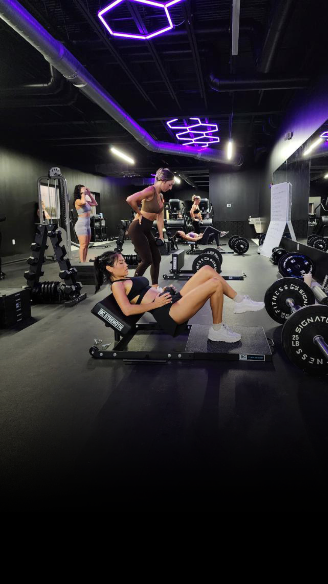 A woman doing hip thrust exercises in the gym with weights on a specialized bench, others exercising nearby.